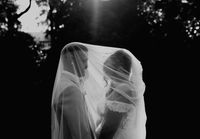 Black and white image of a bride and groom under a veil holding hands looking at each other.